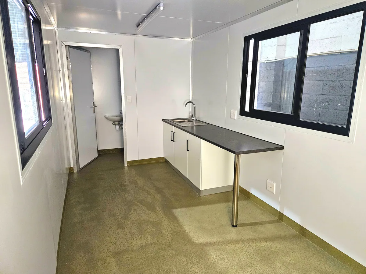 Container canteen interior with a stainless-topped counter, white cabinets, wash basin and two black-framed windows