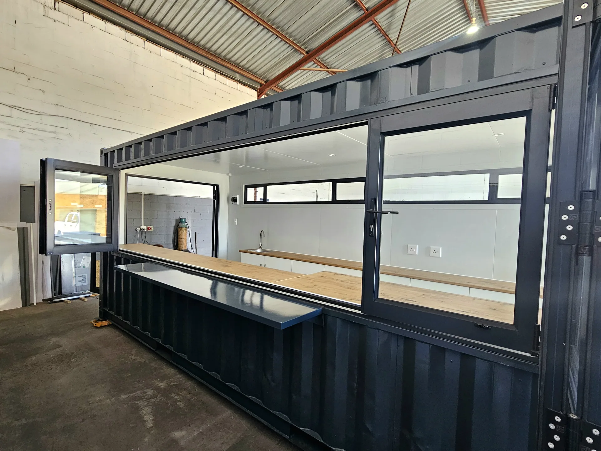 Full side view of a dark container coffee shop with a raised serving hatch and long timber bar counter under a steel roof