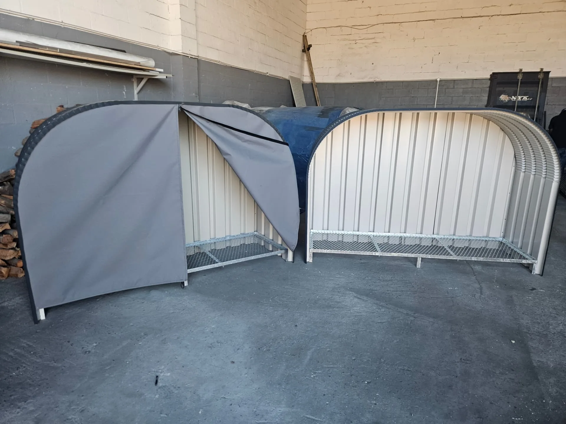 Two firewood sheds in the workshop, one with a grey canvas cover partly open and one open-fronted with a cream back panel