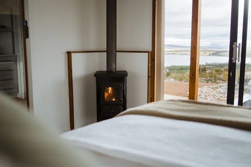 Container house bedroom with a made bed, compact wood-burning stove and glazed door opening towards the landscape