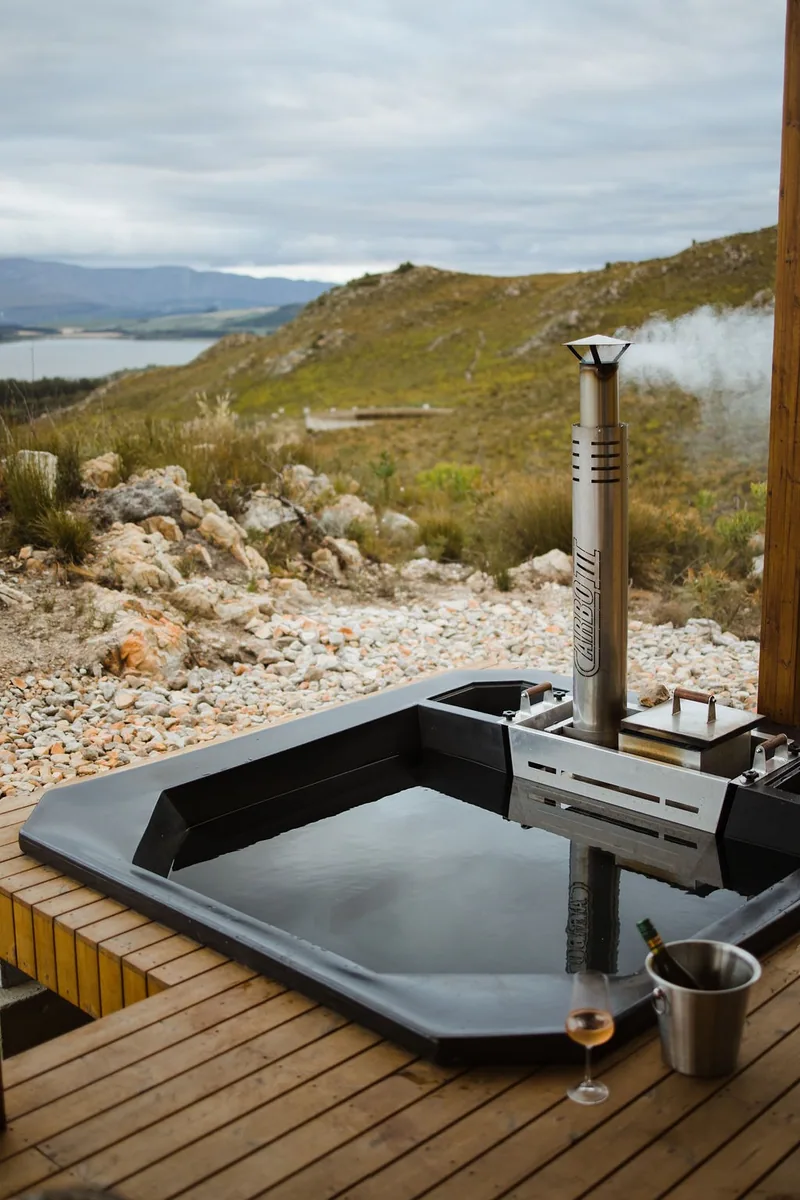 Outdoor wood-fired hot tub with a steel chimney on a timber deck, with mountains behind