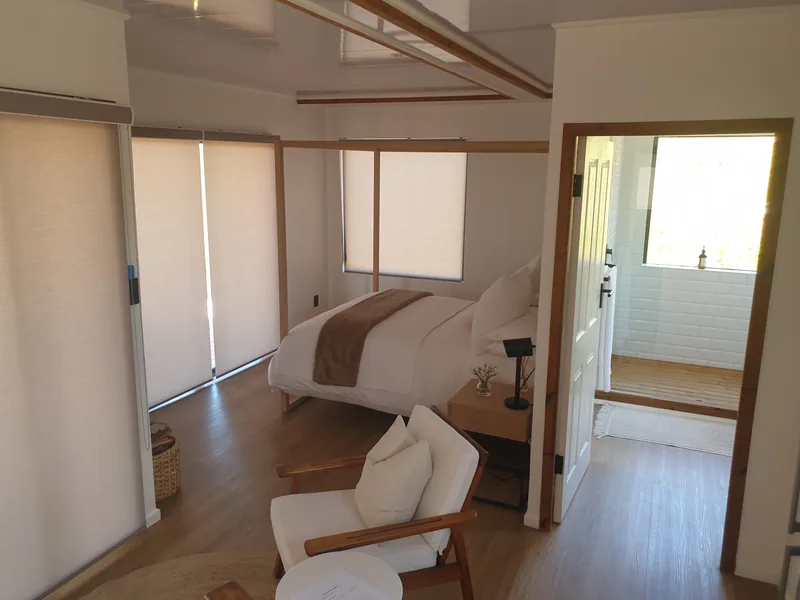Container house bedroom with a made bed, armchair and blinds, with the bathroom visible beyond