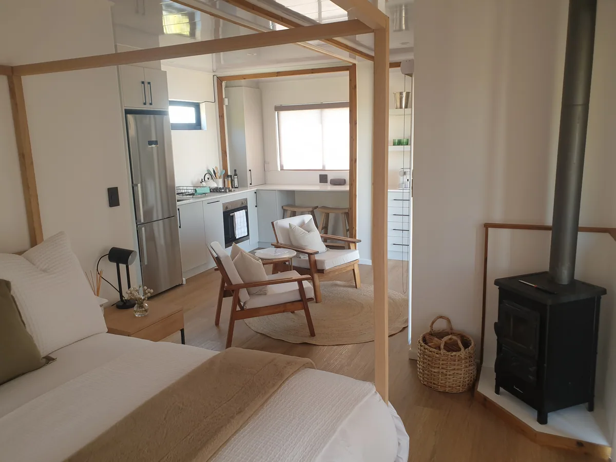 Container house interior showing a bed in the foreground, wood-burning stove and open-plan kitchen and seating area