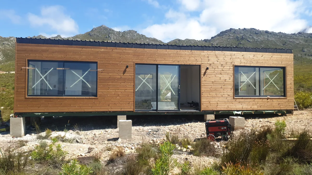 Long timber-clad modular home with a run of black-framed windows, standing in open veld below mountains