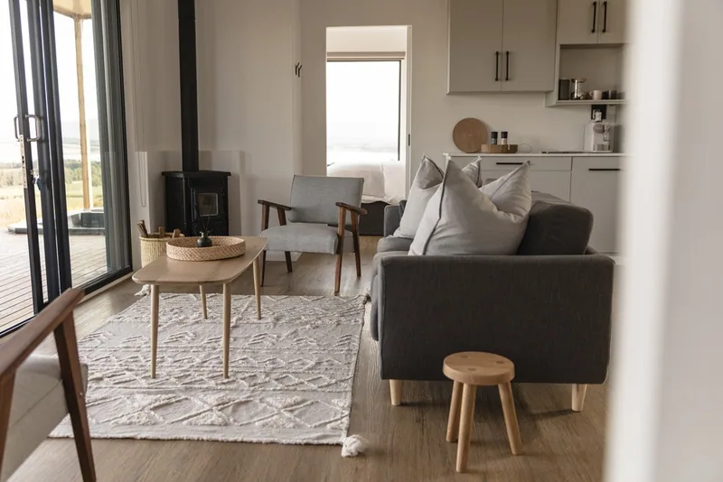 Open-plan living area with a sofa, timber coffee table, rug and a wood-burning stove beside glazed doors