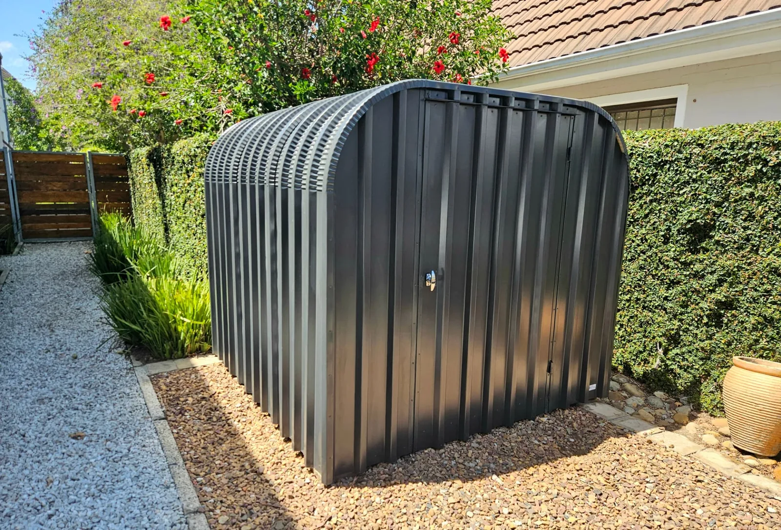 Completed charcoal bike shed installed on a gravel bed beside a garden path, with hedging and flowering shrubs in the background