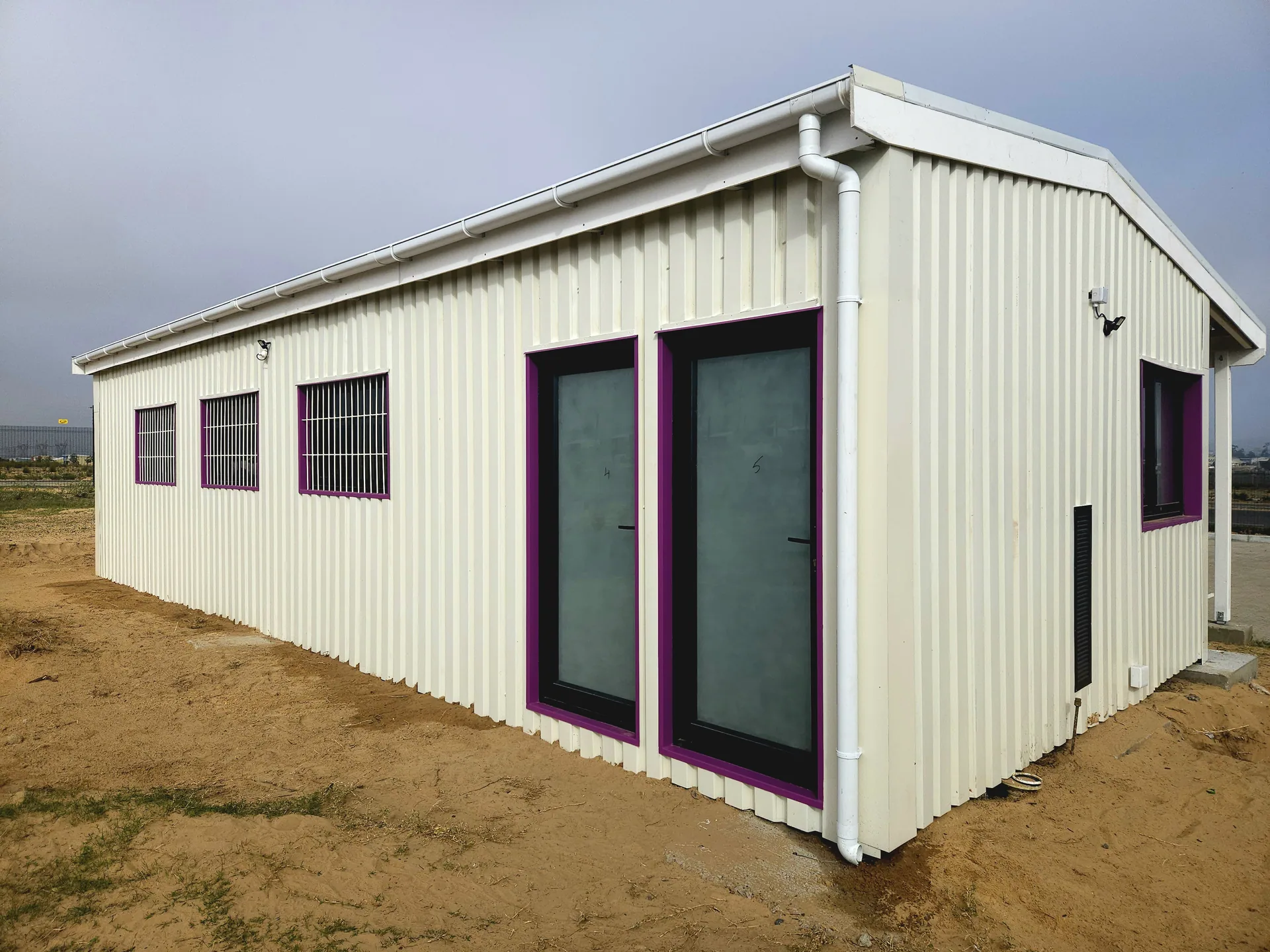 Angled exterior of a white IBR-clad container classroom with pink-framed doors and windows under a pitched roof