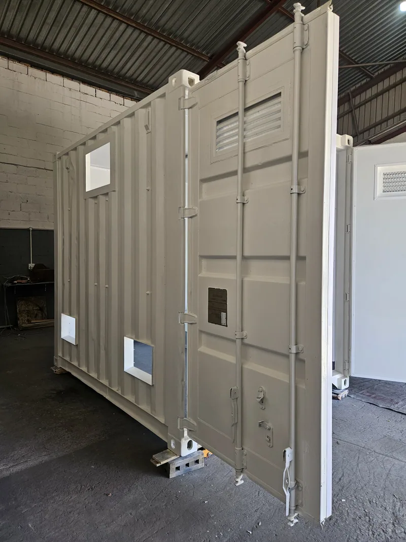 Exterior of a cream painted short container standing on timber blocks inside a workshop, doors open with louvred vents fitted