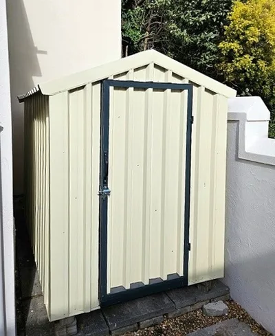 Cream garden tool shed with a dark-framed lockable door, installed on a paved base beside a wall