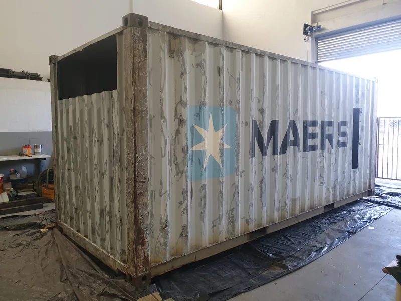 Weathered shipping container with third-party shipping line branding standing on plastic sheeting in a workshop