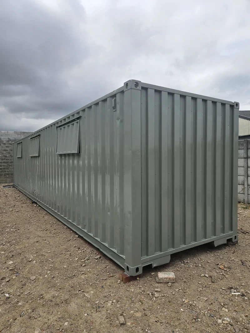 Pale green container standing on timber blocks on a sandy site under a cloudy sky