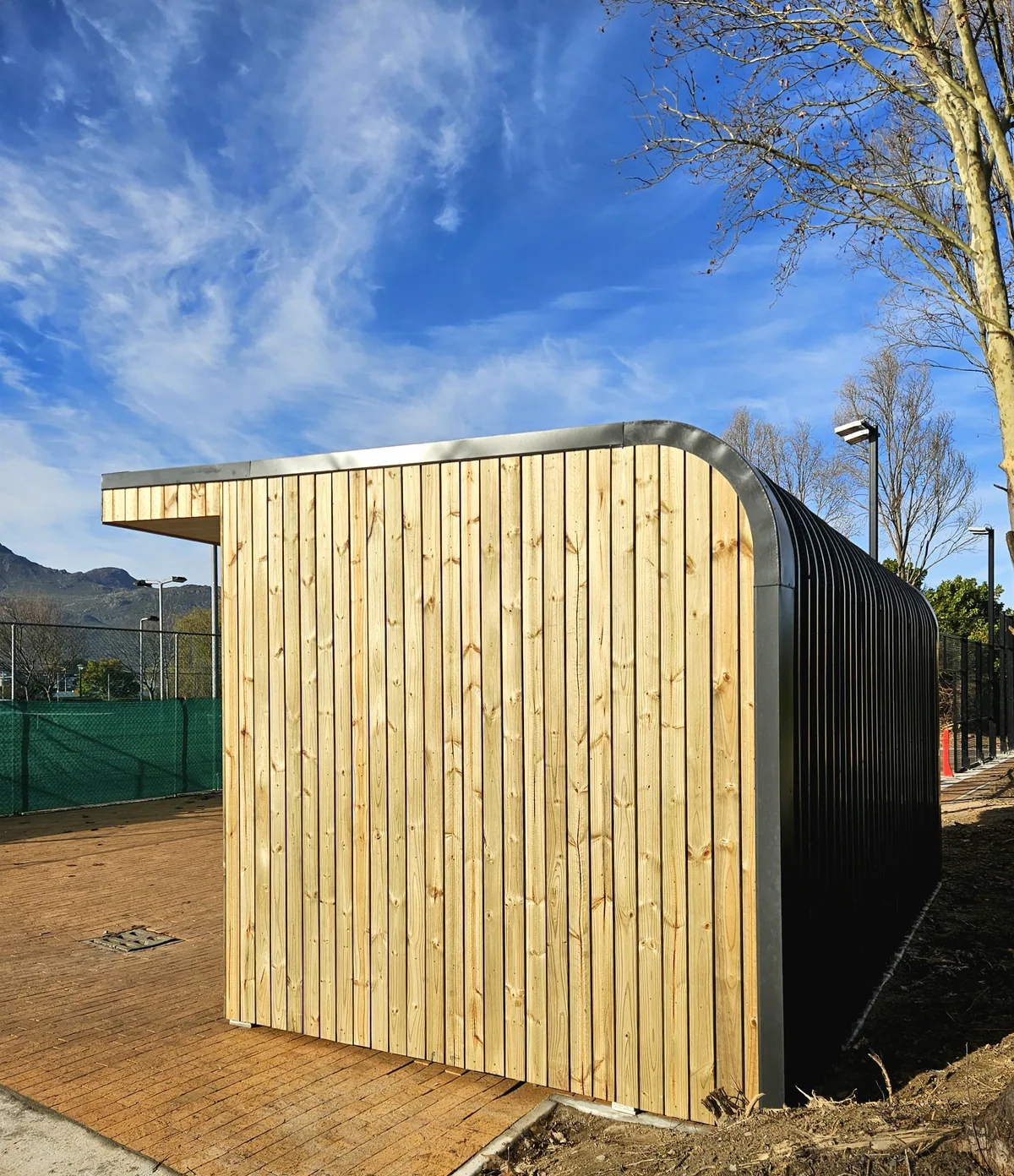 Rear of a timber-clad modular unit with a curved corrugated steel wall, beside a fenced court
