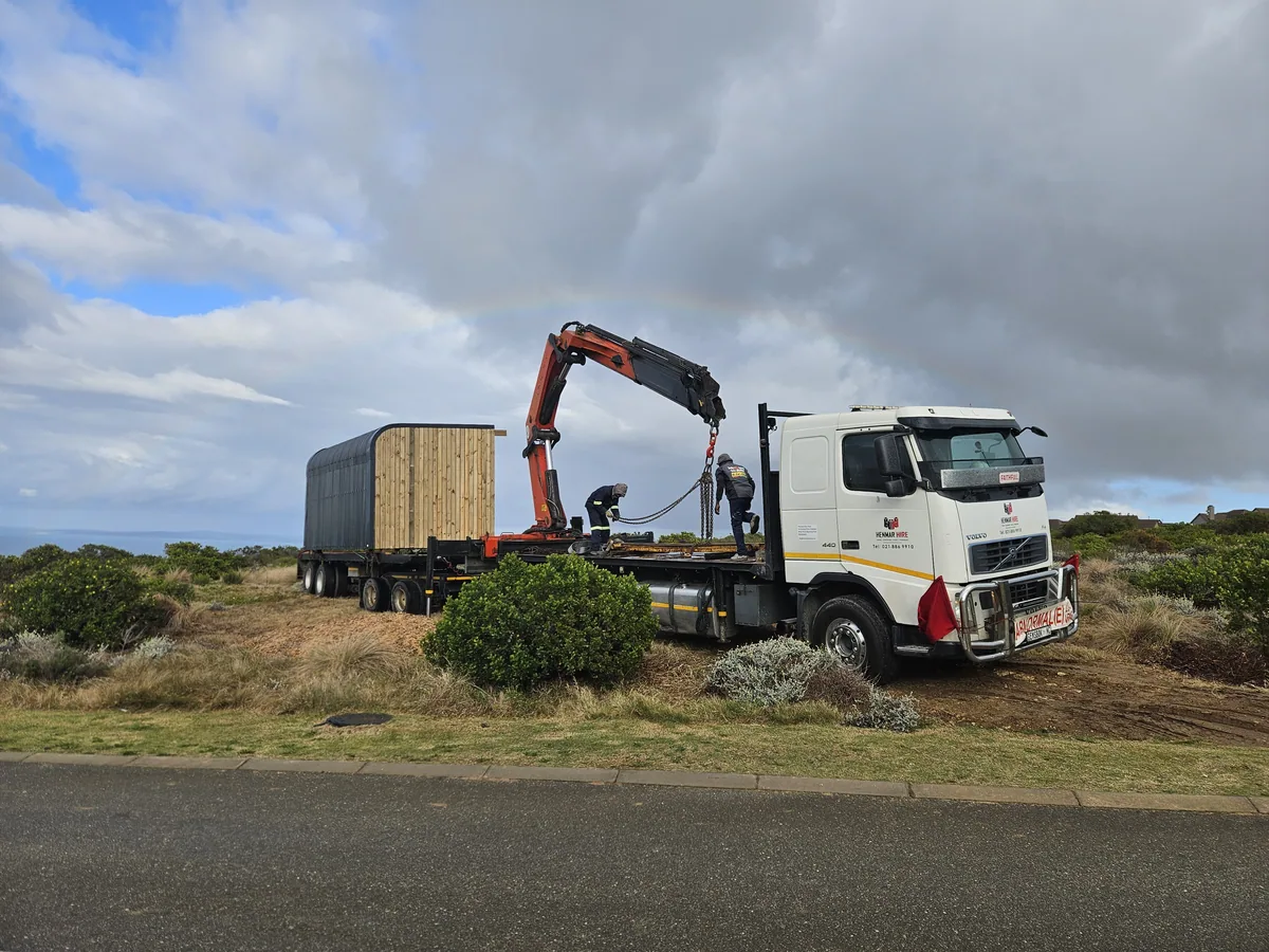 Truck-mounted crane and flatbed truck carrying a wrapped modular unit on a roadside verge