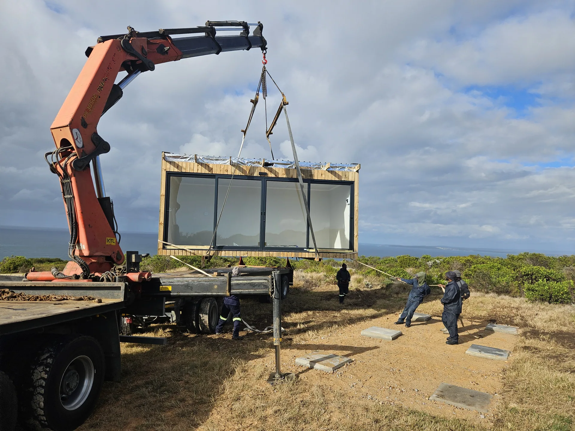 Red truck-mounted crane lowering a glazed timber-clad pod onto foundation pads on a coastal site, with workers guiding it