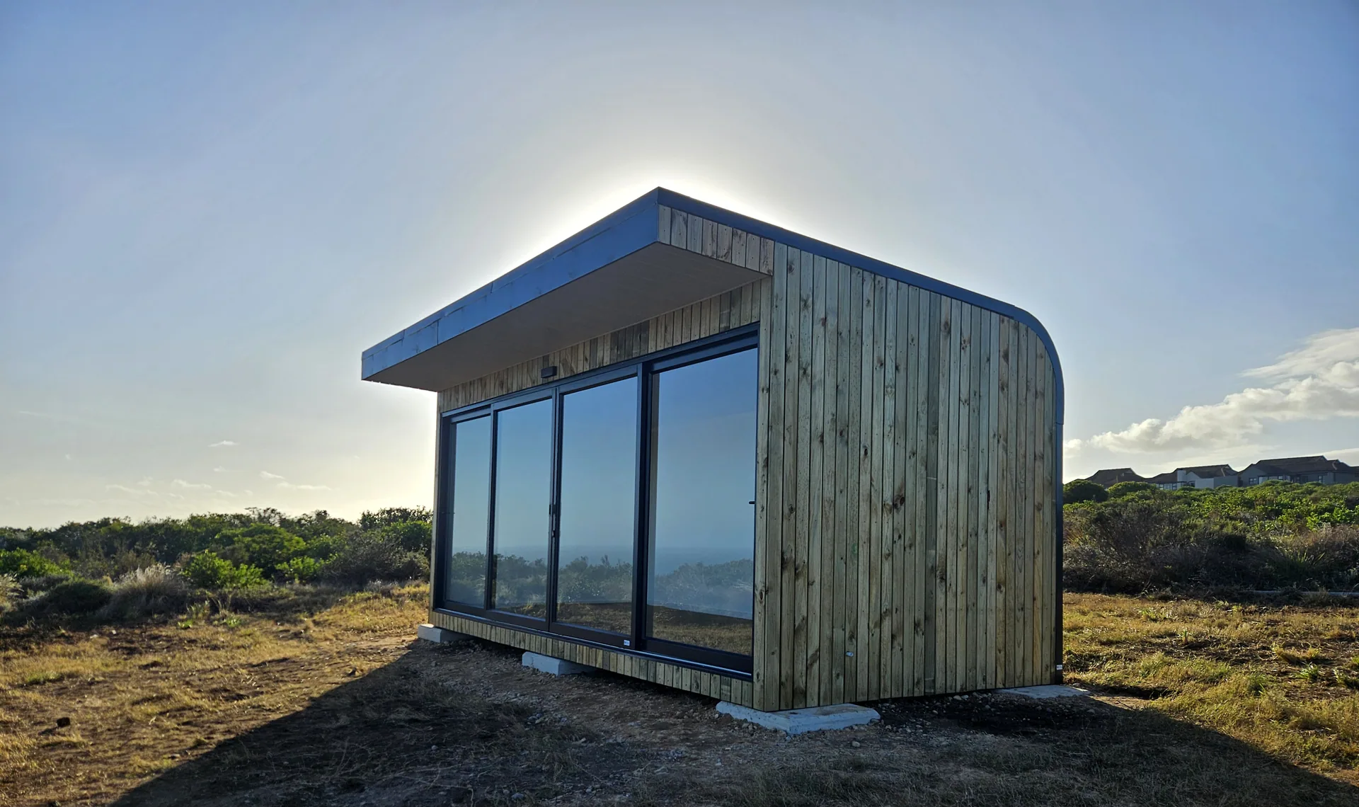 Timber-clad pod with a curved roof and full-height glazing standing on concrete pads in coastal fynbos