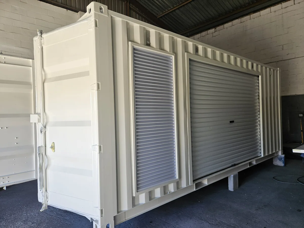 White container with an open personnel door and an external roller shutter, standing on blocks in a workshop